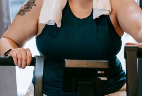 Recall Training - Smiling plus size female with towel exercising on cross trainer machine while looking at camera during workout in fitness studio