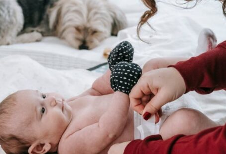 Dog Scratching - Crop anonymous mother putting on diaper on adorable infant baby wearing anti scratching mittens while lying on comfortable couch near hairy dog in light room